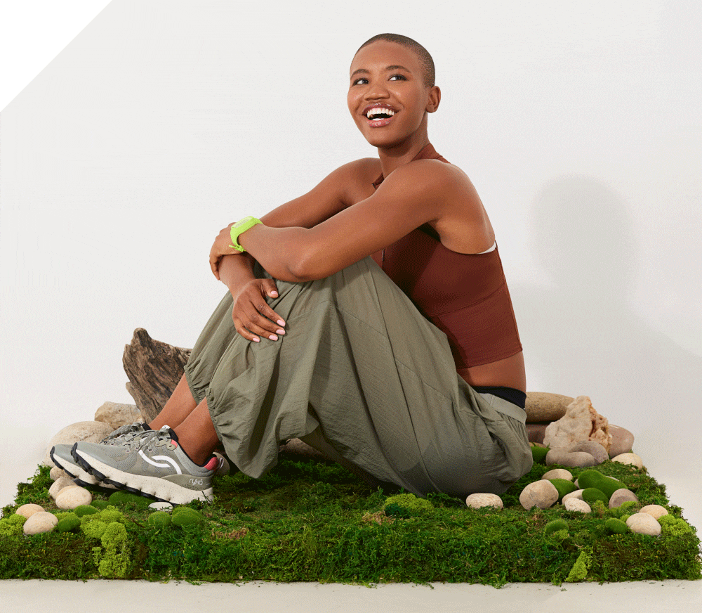 Woman sitting in the grass wearing a pair of gray walking sneakers with a white sole. The image then transitions to a closeup of the shoes. 
