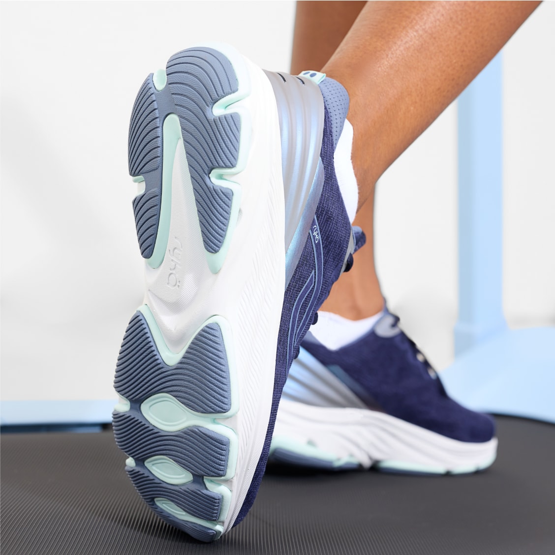 A woman wearing a pair of blue walking sneakers with one foot placed vertically, highlighting the tread on the sole. 