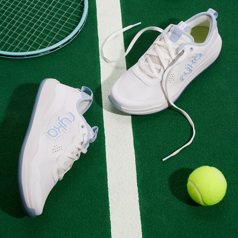 A pair of Rykä court agility shoes on a tennis court next to a tennis racket and a tennis ball