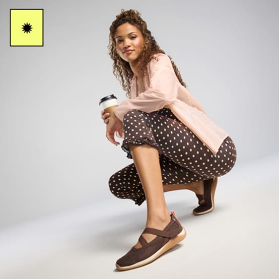 Woman kneeling down wearing a pink jacket, brown polka dot leggings, and brown Mary Jane sneakers. 