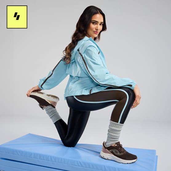 Woman kneeling on a workout mat wearing a blue jacket, black leggings, and brown sneakers. 