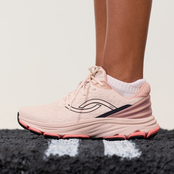 A woman standing on an asphalt surface wearing a pair of light pink sneakers with black and coral accents. 