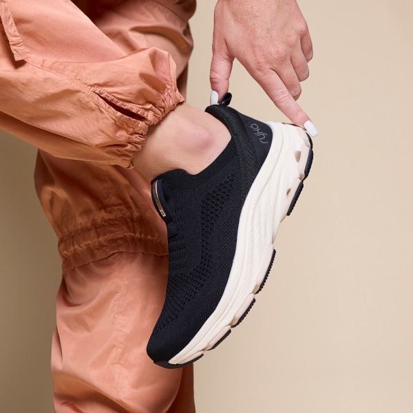 A woman standing up wearing peach satin pants and a pair of women's slip on sneakers in a black knit material. 