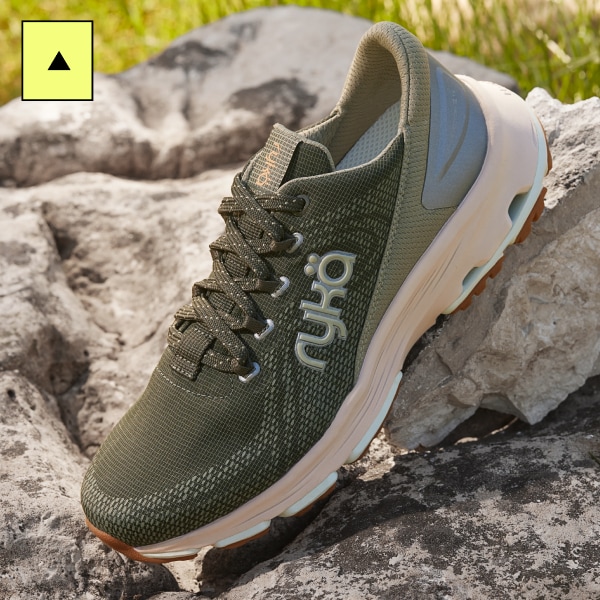 A dark green woman's walking sneaker with an off-white sole displayed on a rocky background. 
