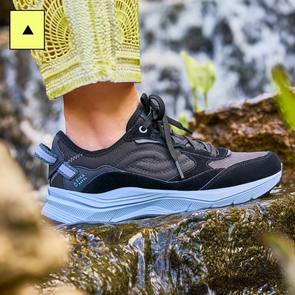 A woman wearing a pair of black and gray trail sneakers with a blue sole against a rocky background. 