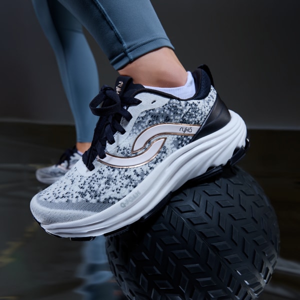 A woman standing with one foot on a medicine ball wearing a pair of blue speckled walking sneakers with a blue sole and black laces. 