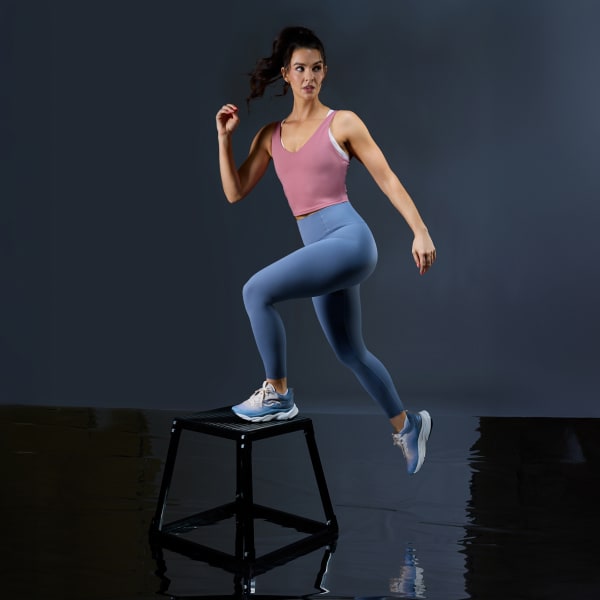 A woman standing with one foot on a stool wearing a pink tank, blue leggings, and a pair of blue and white walking sneakers. 
