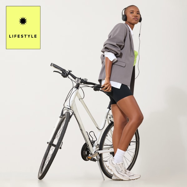 A woman leaning on a bike wearing a gray blazer, black shorts, and chunky white sneakers. A yellow graphic says 