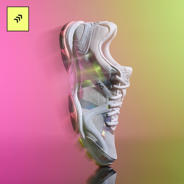A grey and silver women's training sneaker displayed vertically on a pink and yellow gradient background. 
