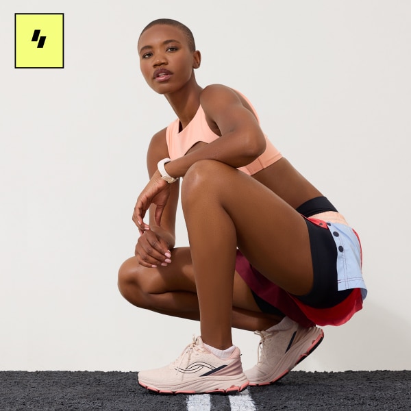 A woman stooped down wearing a peach sports bra, blue and red shorts, and a pair of pink walking sneakers.  